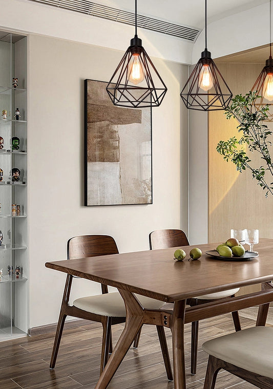 Aten - Suspension - Fer - Noir: Three black geometric-cage pendant lights with exposed Edison bulbs above a wooden dining table, enhancing a modern, minimalist aesthetic.